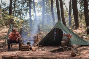 Man enjoying a warm cup of drink by his camp in the middle of the woods.