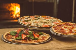 Three pizzas on pans with a brick oven ablaze in the background.